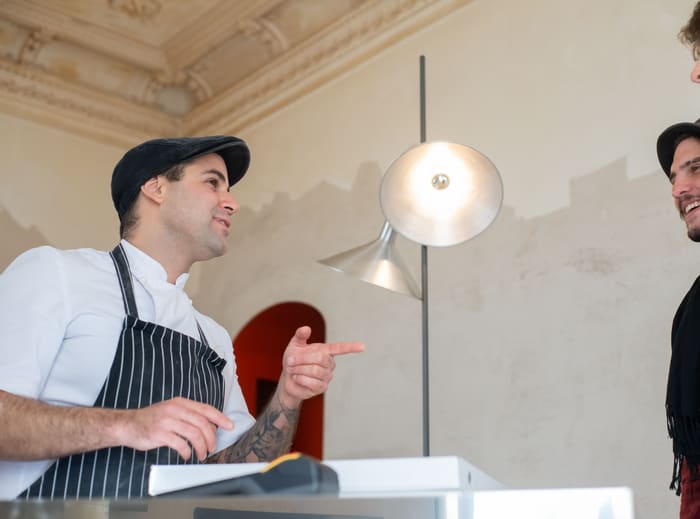 A cafe owner in an apron talking with a customer across the counter